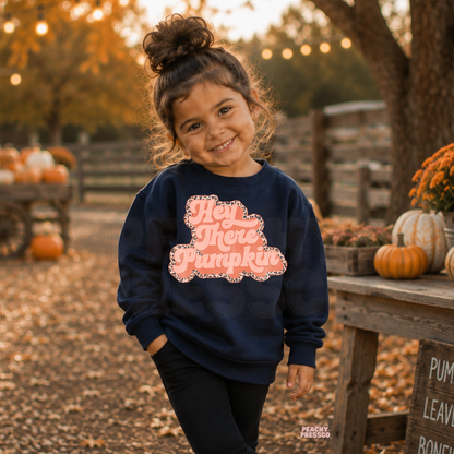 Cute toddler girl wearing a navy "Hey There Pumpkin" sweatshirt with a retro pink fall design at a pumpkin patch with pumpkins and autumn décor.