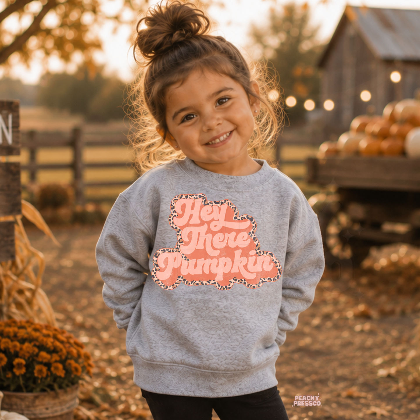 Toddler girl smiling in a sport grey "Hey There Pumpkin" sweatshirt with a pink leopard print retro graphic at a fall pumpkin patch.