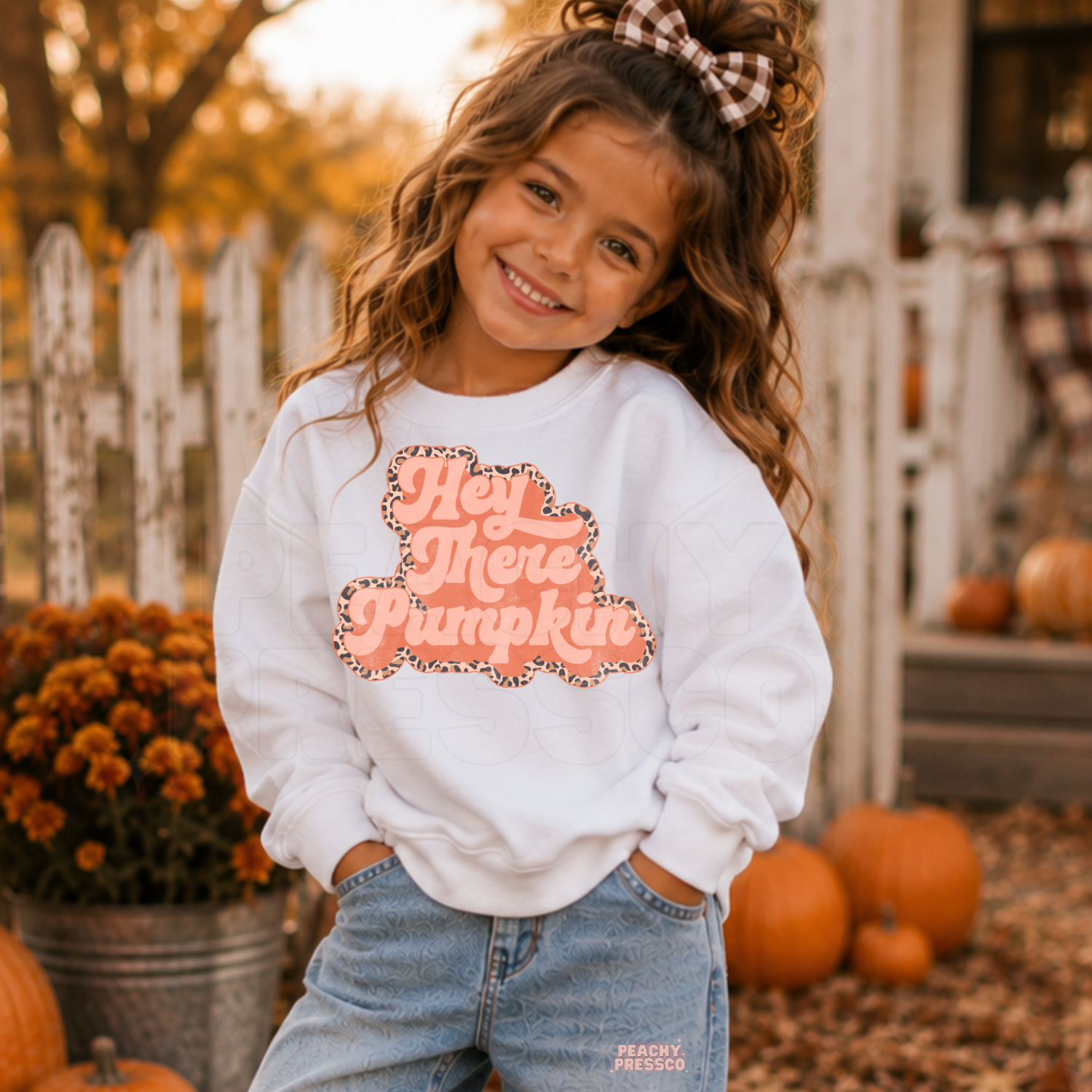 Smiling toddler girl wearing a white "Hey There Pumpkin" sweatshirt with a pink retro design at a cozy pumpkin patch in autumn.