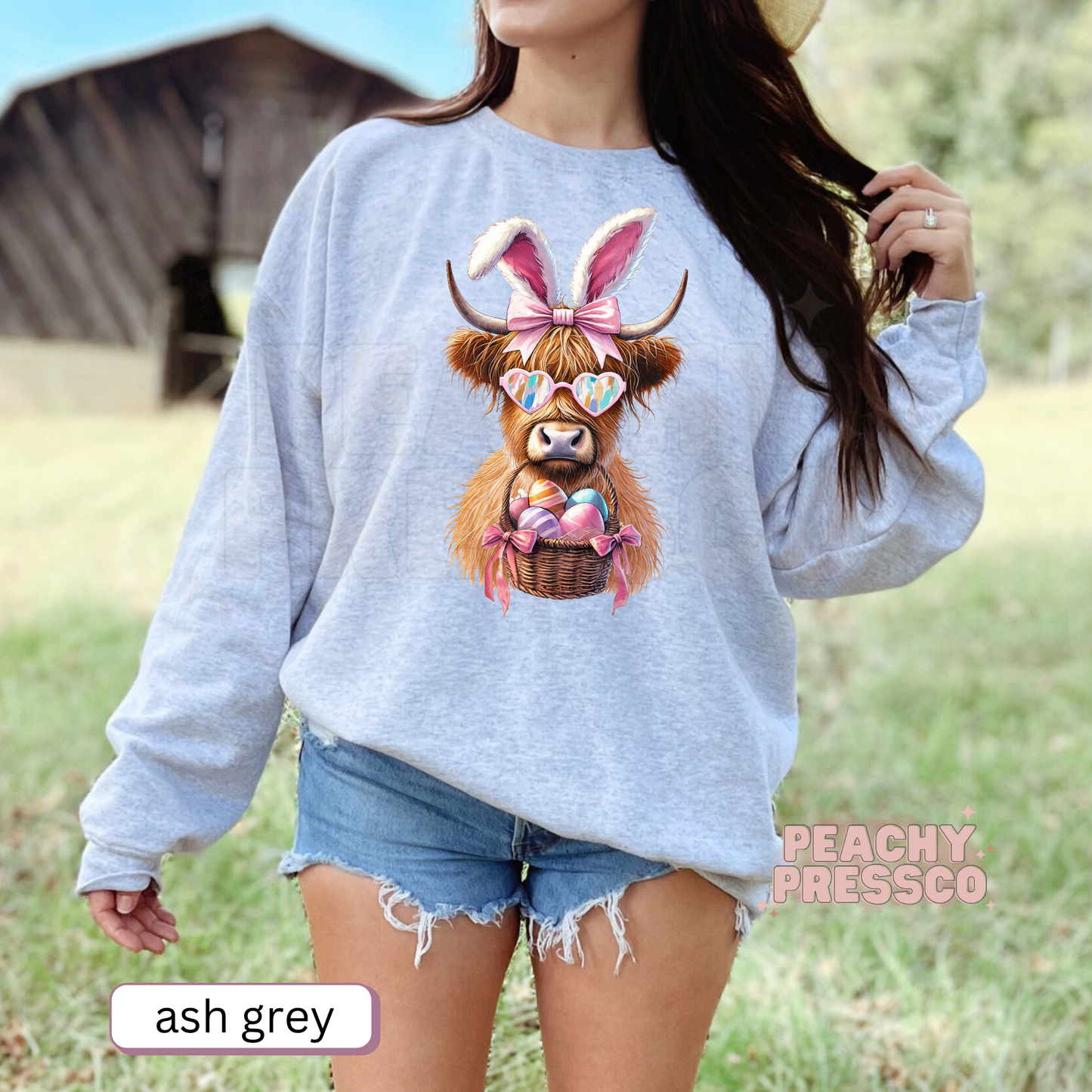Woman wearing an ash grey crewneck sweatshirt featuring a Highland cow dressed as the Easter Bunny with heart sunglasses and a basket of colorful Easter eggs.