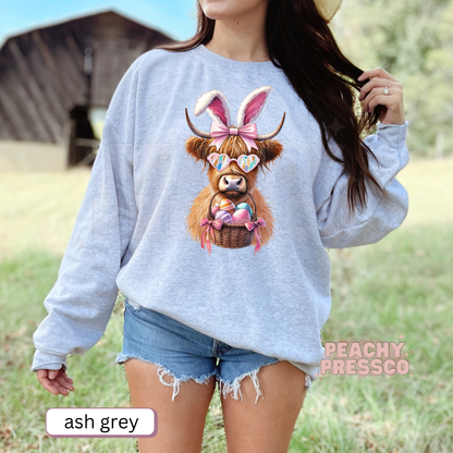 Woman wearing an ash grey crewneck sweatshirt featuring a Highland cow dressed as the Easter Bunny with heart sunglasses and a basket of colorful Easter eggs.