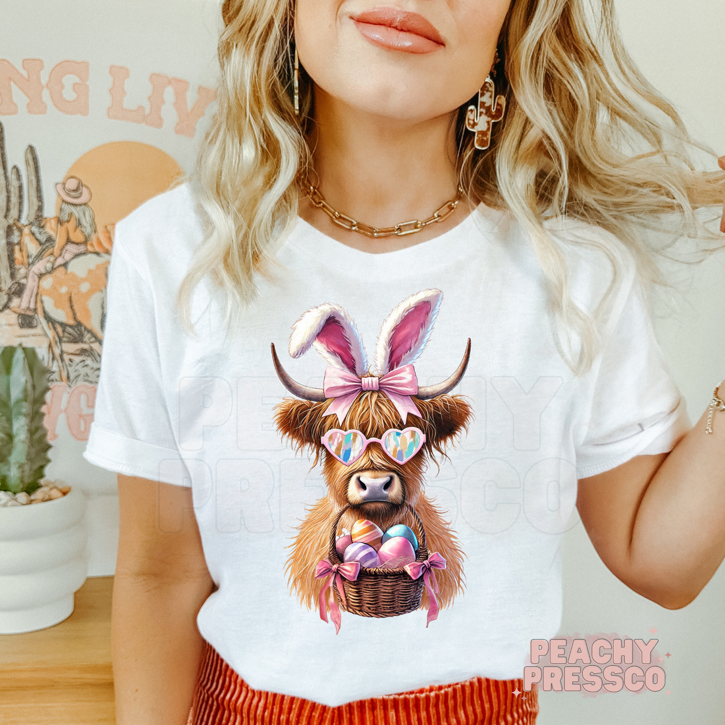 Woman wearing a white t-shirt featuring a cute Highland cow Easter graphic with bunny ears, pink bow, heart sunglasses, and a basket of pastel Easter eggs.