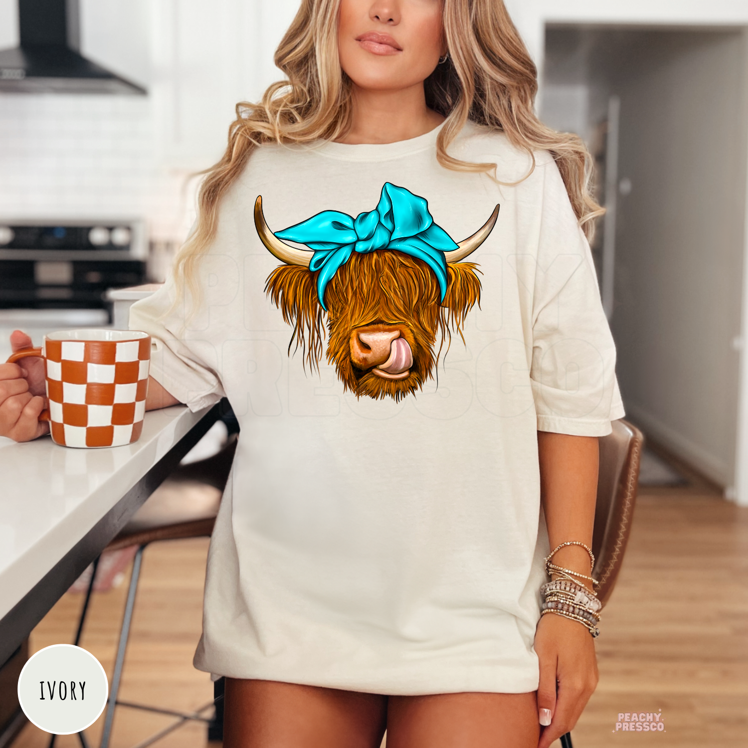 Woman wearing an ivory t-shirt with a cute Highland cow graphic wearing a turquoise bow and playful expression.