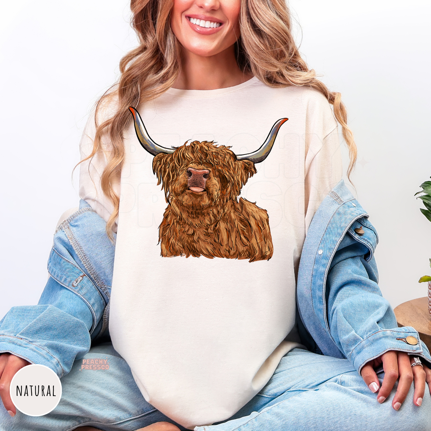 Woman wearing a natural colored shirt with a Highland cow graphic, rustic farmhouse apparel for western style lovers.