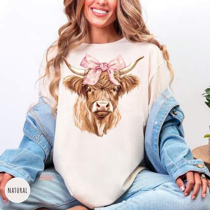 Woman wearing a natural sweatshirt featuring a fluffy Highland cow with a pink bow and western rustic country aesthetic.