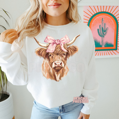 Woman wearing a white sweatshirt featuring a cute Highland cow with a pink bow, western farmhouse inspired graphic.