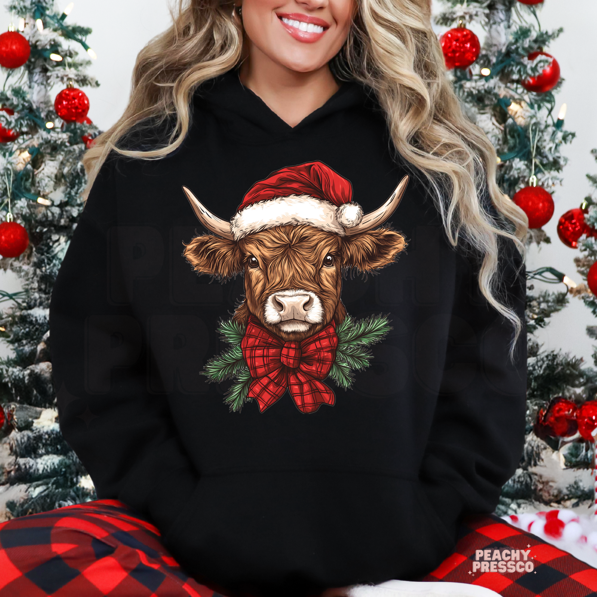 A woman wearing a black hoodie with a graphic of a brown Scottish Highland cow wearing a red Santa hat and red plaid bow tie, surrounded by green pine branches. She is sitting in front of decorated Christmas trees, wearing red and black plaid pants.