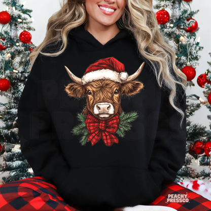 A woman wearing a black hoodie with a graphic of a brown Scottish Highland cow wearing a red Santa hat and red plaid bow tie, surrounded by green pine branches. She is sitting in front of decorated Christmas trees, wearing red and black plaid pants.