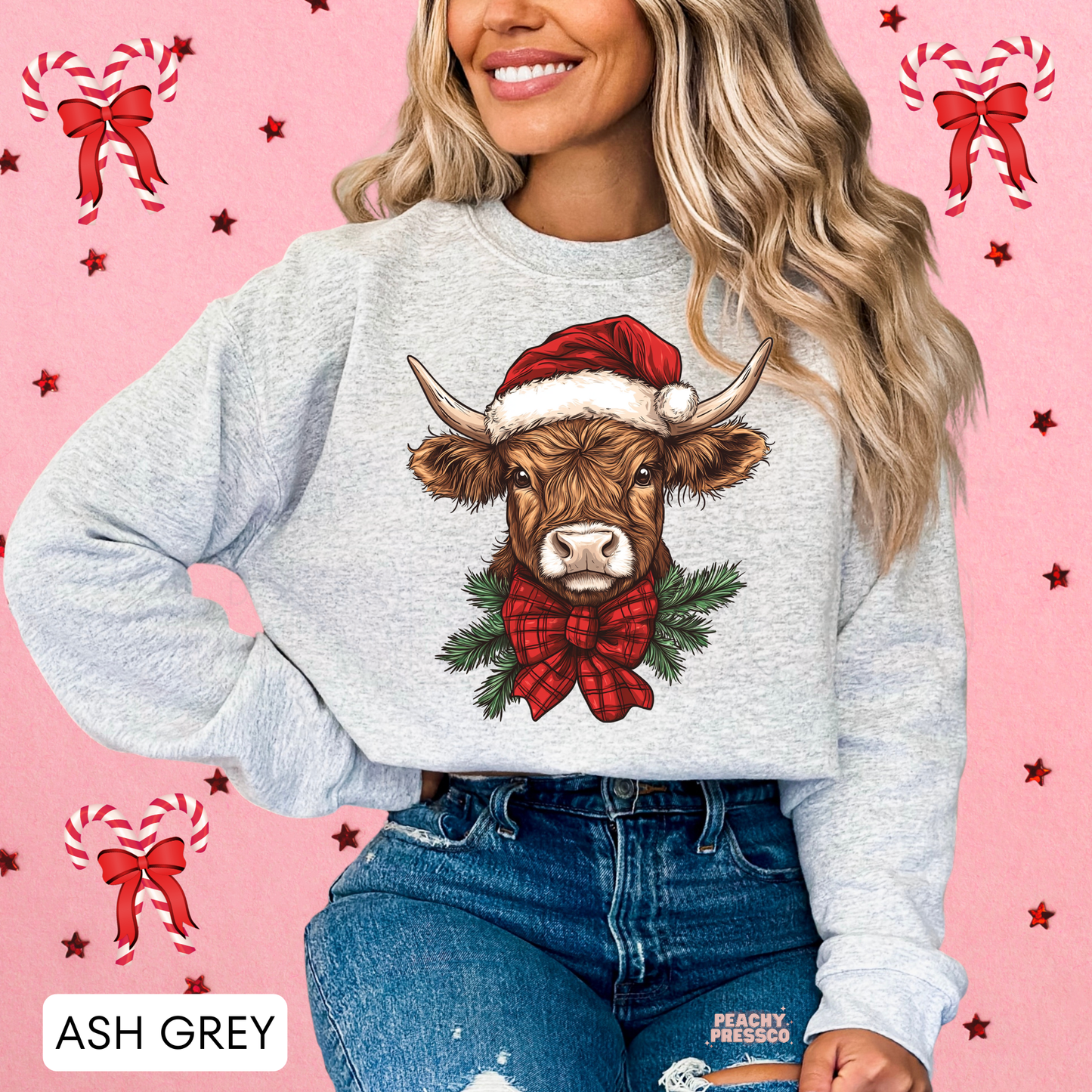A woman wearing a ash grey crewneck sweatshirt with a graphic of a Highland cow in a Santa hat and red bow, paired with blue jeans, set against a pink background with holiday bow illustrations.
