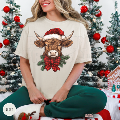 Woman sitting wearing an ivory Christmas t-shirt printed with a Highland cow wearing a Santa hat and plaid bow, paired with green sweatpants.