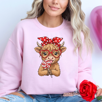Light pink sweatshirt with a charming Highland cow in heart glasses, a red bow, and a candy swirl lollipop for a festive Valentine's look.