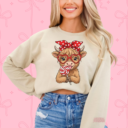 Sand sweatshirt displaying a sweet Highland cow wearing a red polka dot bow and heart glasses while holding a Valentine's lollipop.