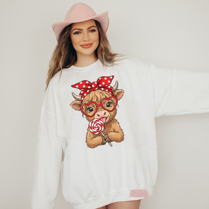 White sweatshirt featuring a cute Highland cow wearing heart-shaped glasses, a red polka dot bow, and holding a heart swirl lollipop for Valentine's Day.