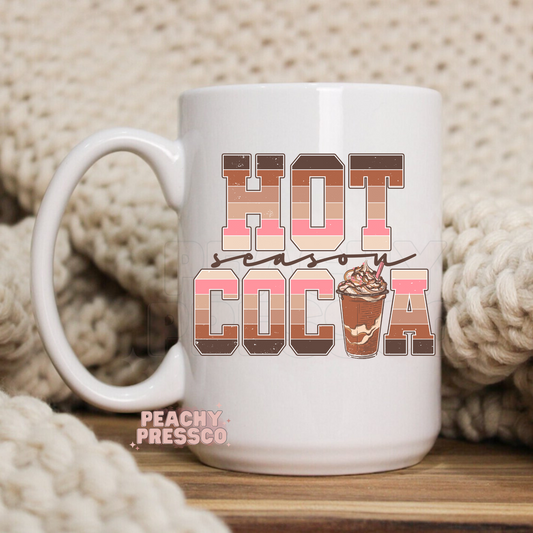 White ceramic coffee mug featuring retro multi-toned block lettering reading Hot Cocoa Season with an iced cocoa cup illustration.