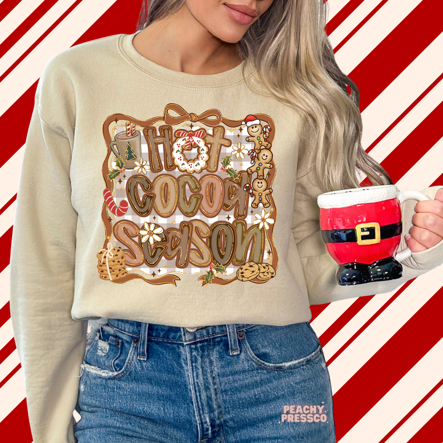 A woman wearing a beige crewneck sweatshirt with a 'Hot Cocoa Season' graphic, holding a red and white Santa mug against a red and white diagonal striped background.