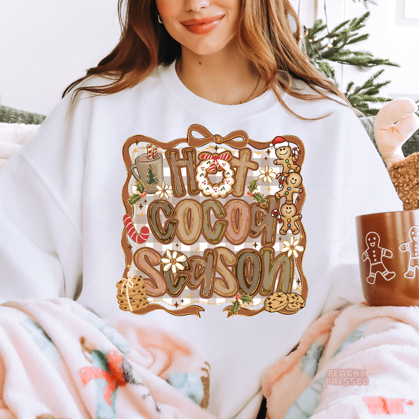Woman relaxing on a couch under a fuzzy blanket with a gingerbread mug, wearing a white Hot Cocoa Season sweatshirt.