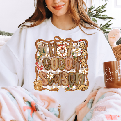 Woman relaxing on a couch under a fuzzy blanket with a gingerbread mug, wearing a white Hot Cocoa Season sweatshirt.