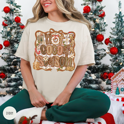 A woman wearing a ivory colored graphic t-shirt that reads 'Hot Cocoa Season' in a festive gingerbread-themed design, paired with dark green sweatpants, sitting in front of decorated Christmas trees.