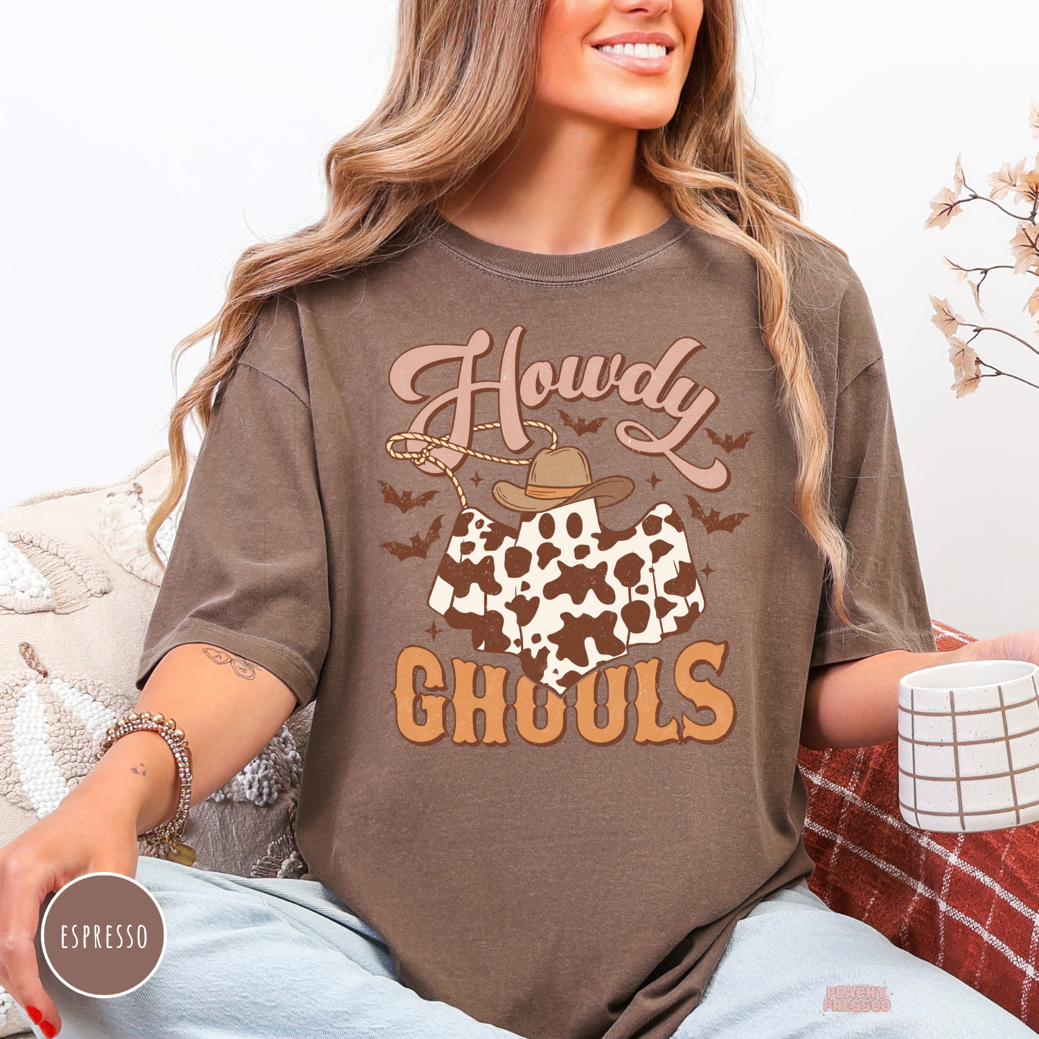 Woman wearing an espresso Comfort Colors t-shirt featuring a Howdy Ghouls western ghost graphic with cow print, cowboy hat, bats and spooky fall design.