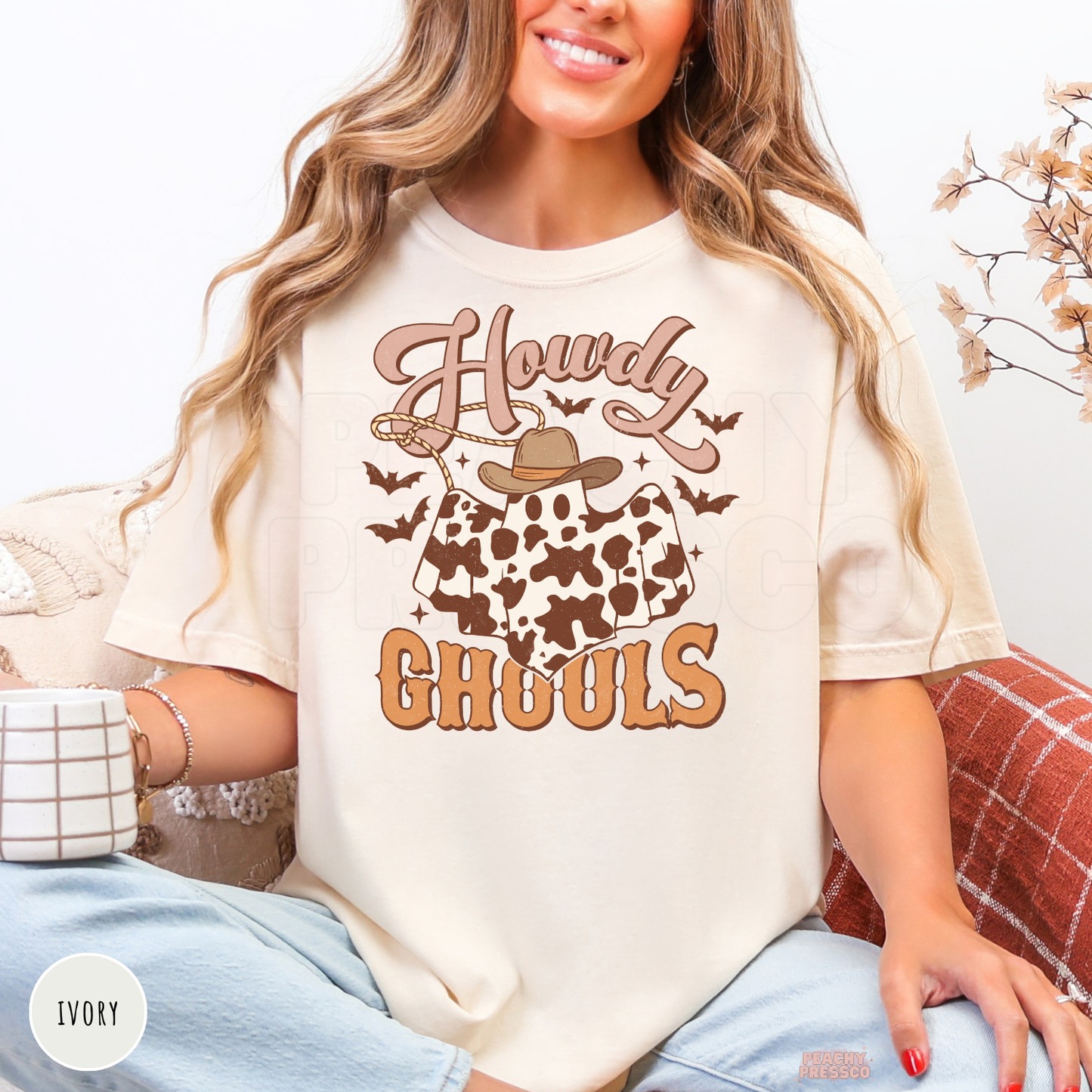 Woman wearing an ivory Comfort Colors t-shirt with a western Howdy Ghouls ghost graphic featuring cow print, cowboy hat and spooky bats.