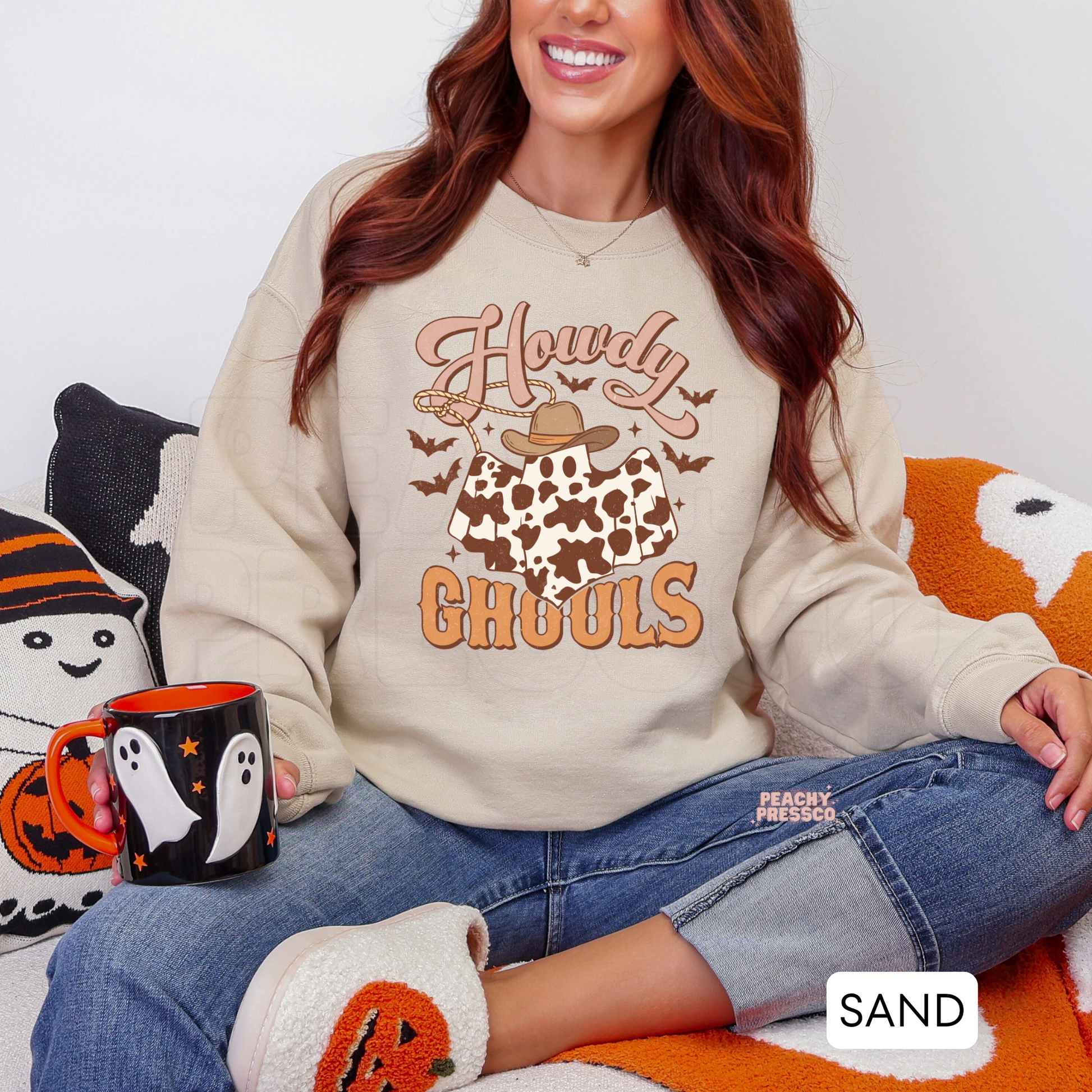 Woman wearing a sand crewneck sweatshirt featuring a Howdy Ghouls cowboy ghost graphic with cow print, bats and western Halloween design.