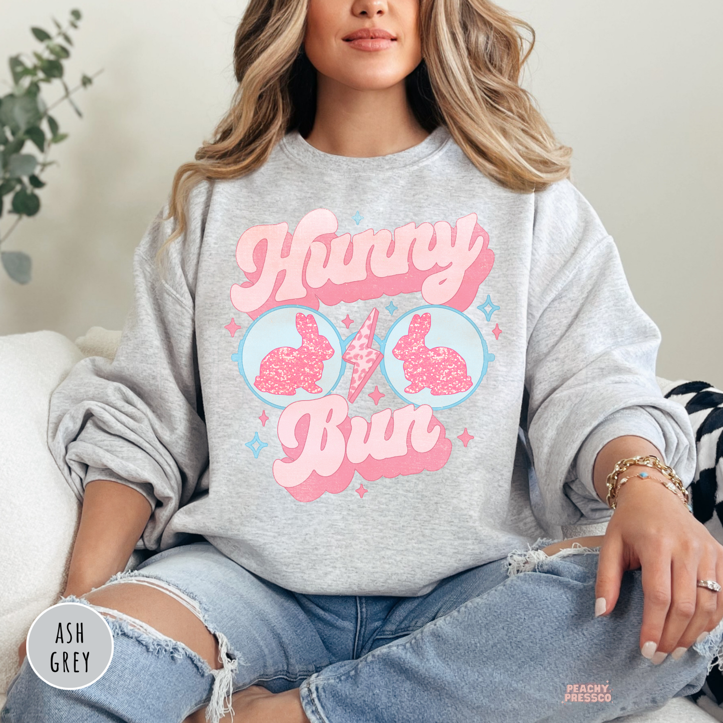 Woman wearing an ash grey sweatshirt with a retro Hunny Bun Easter bunny sunglasses graphic in pink and blue pastel colors.