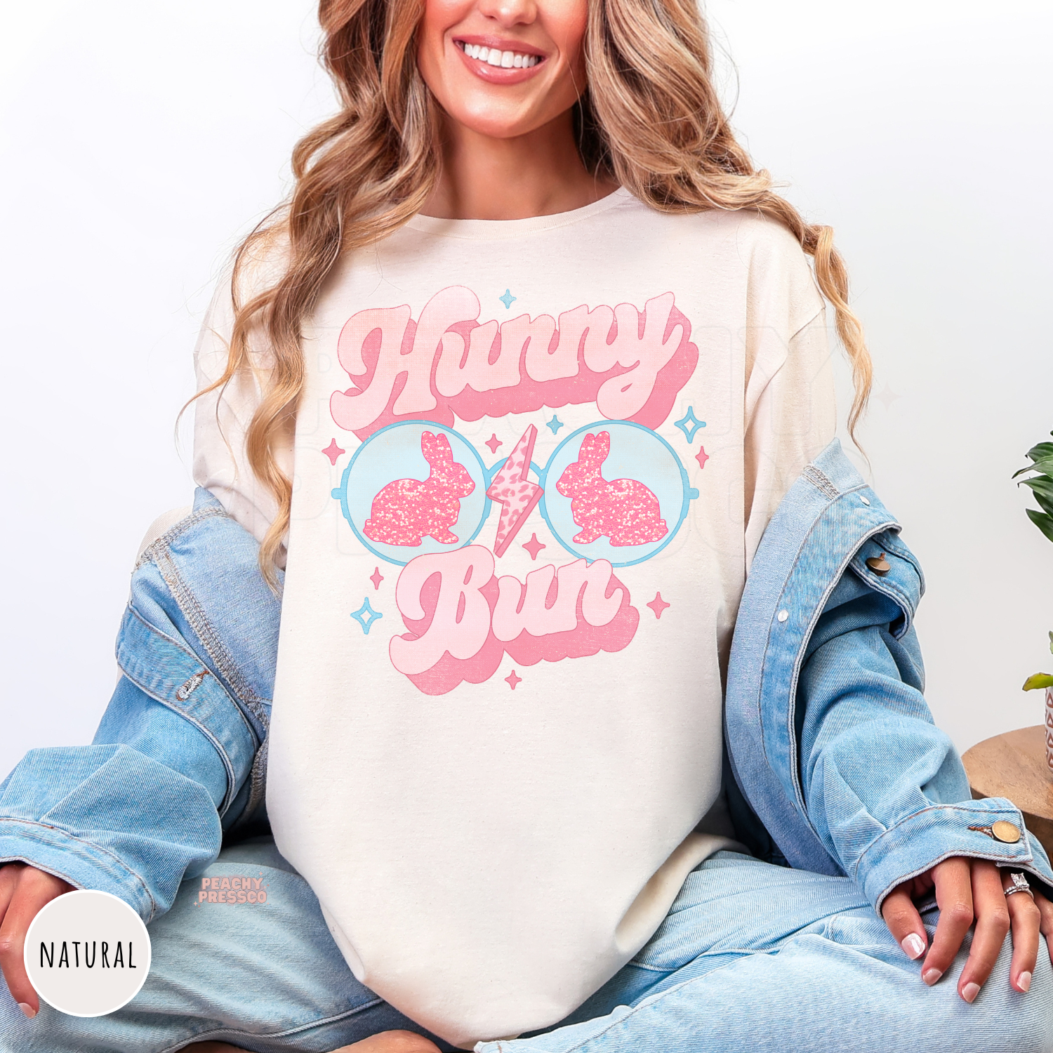 Woman wearing a natural shirt featuring a retro "Hunny Bun" Easter graphic with pink glitter bunny sunglasses and pastel spring colors.