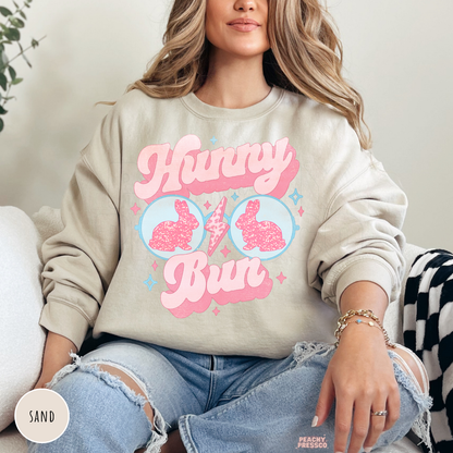 Woman wearing a sand crewneck sweatshirt featuring a cute Hunny Bun Easter bunny sunglasses graphic with retro pastel design.