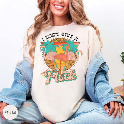 Natural Gildan unisex t-shirt featuring three pink flamingos, palm trees, and a retro sunset with the phrase "I Don't Give a Flock." Vintage tropical graphic tee perfect for summer, beach trips, vacations, pool parties, and flamingo fans.