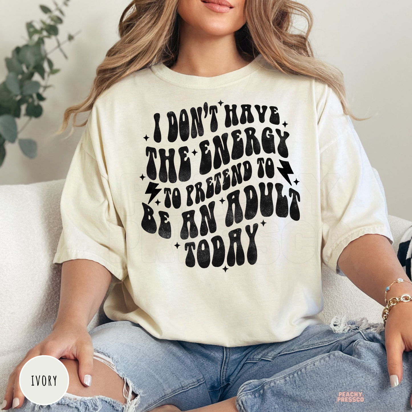 Woman wearing an ivory graphic t-shirt featuring the funny quote "I Don't Have the Energy to Pretend to Be an Adult Today" in retro black lettering.