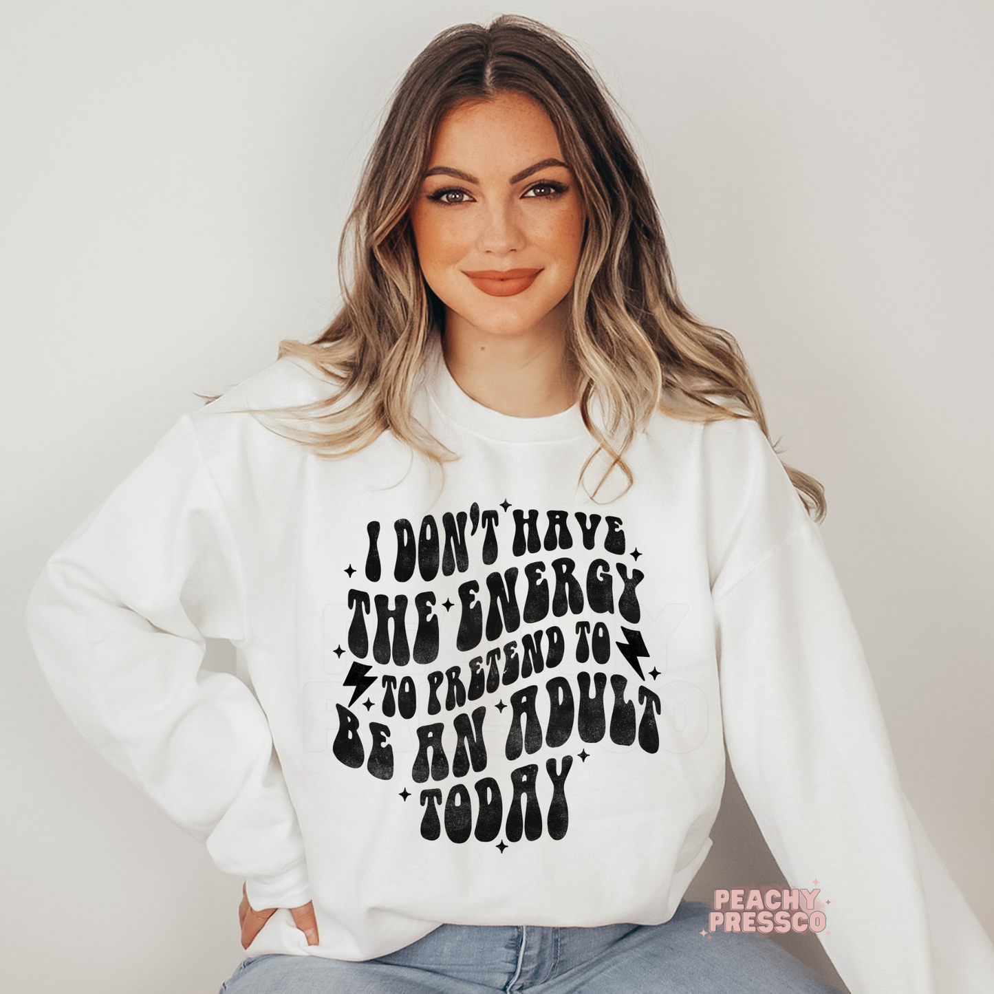 Woman wearing a white graphic sweatshirt with the humorous quote "I Don't Have the Energy to Pretend to Be an Adult Today" in vintage retro typography.
