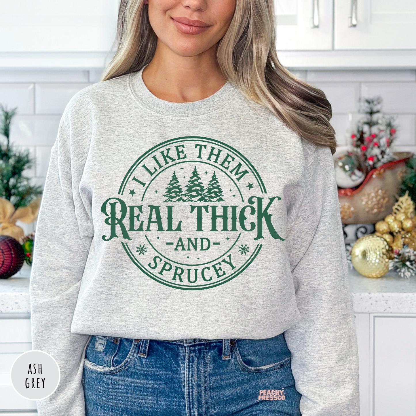 A woman wearing a ash gray crewneck sweatshirt with a dark green circular graphic that reads 'I LIKE THEM REAL THICK AND SPRUCEY' with pine trees in the center. She is wearing blue jeans and standing in a decorated kitchen setting.