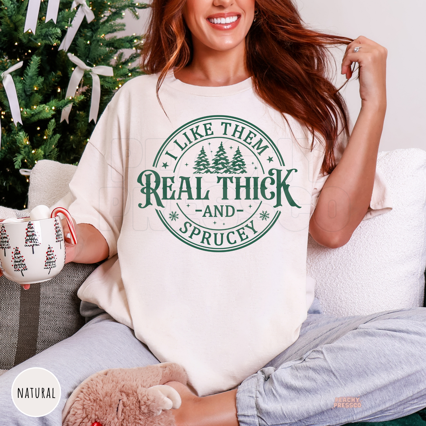 Woman lounging on a couch wearing a natural cream oversized Christmas shirt that says I Like Them Real Thick And Sprucey while holding a mug.