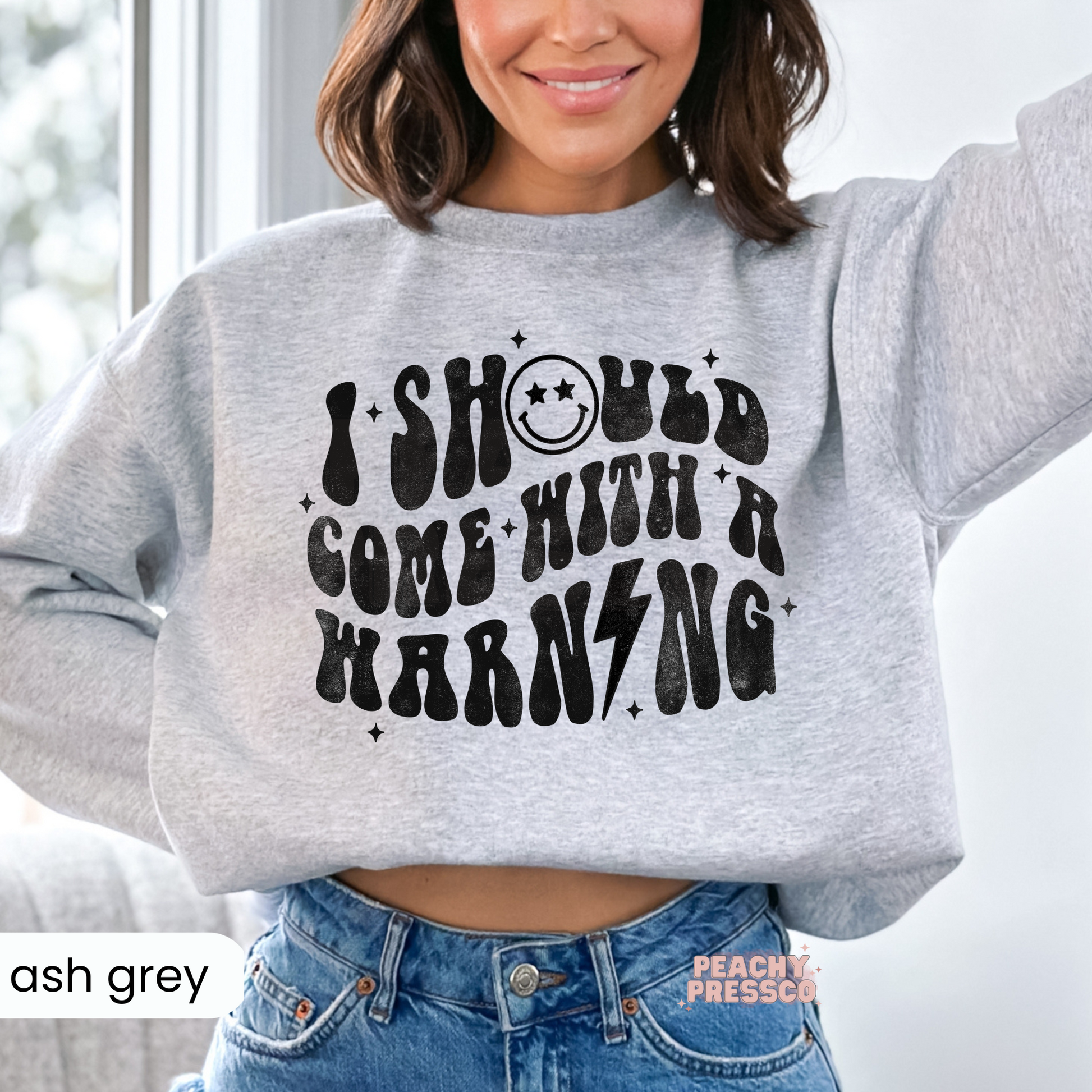 Woman wearing an ash grey cropped sweatshirt featuring the retro "I Should Come With A Warning" graphic and smiley face.