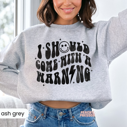 Woman wearing an ash grey cropped sweatshirt featuring the retro "I Should Come With A Warning" graphic and smiley face.