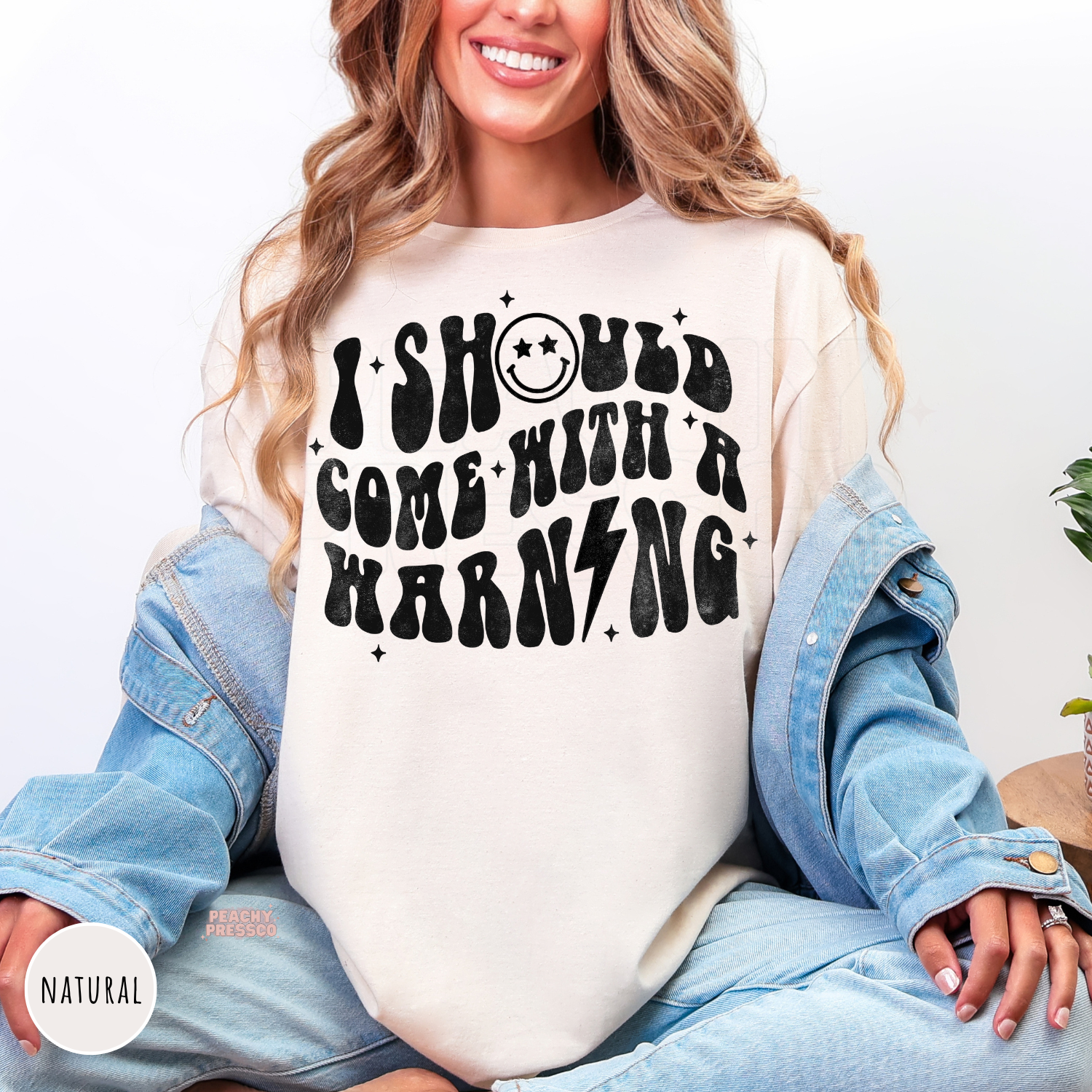 Woman wearing a natural shirt featuring the bold retro "I Should Come With A Warning" graphic.