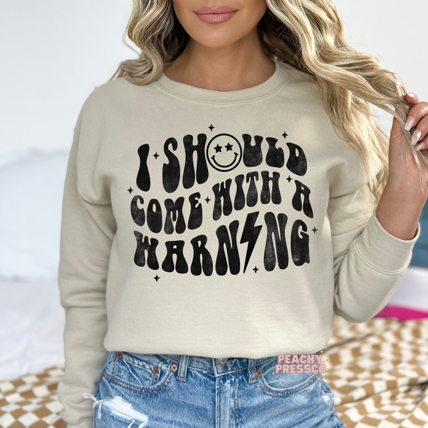 Woman wearing a sand crewneck sweatshirt with a distressed "I Should Come With A Warning" retro graphic.