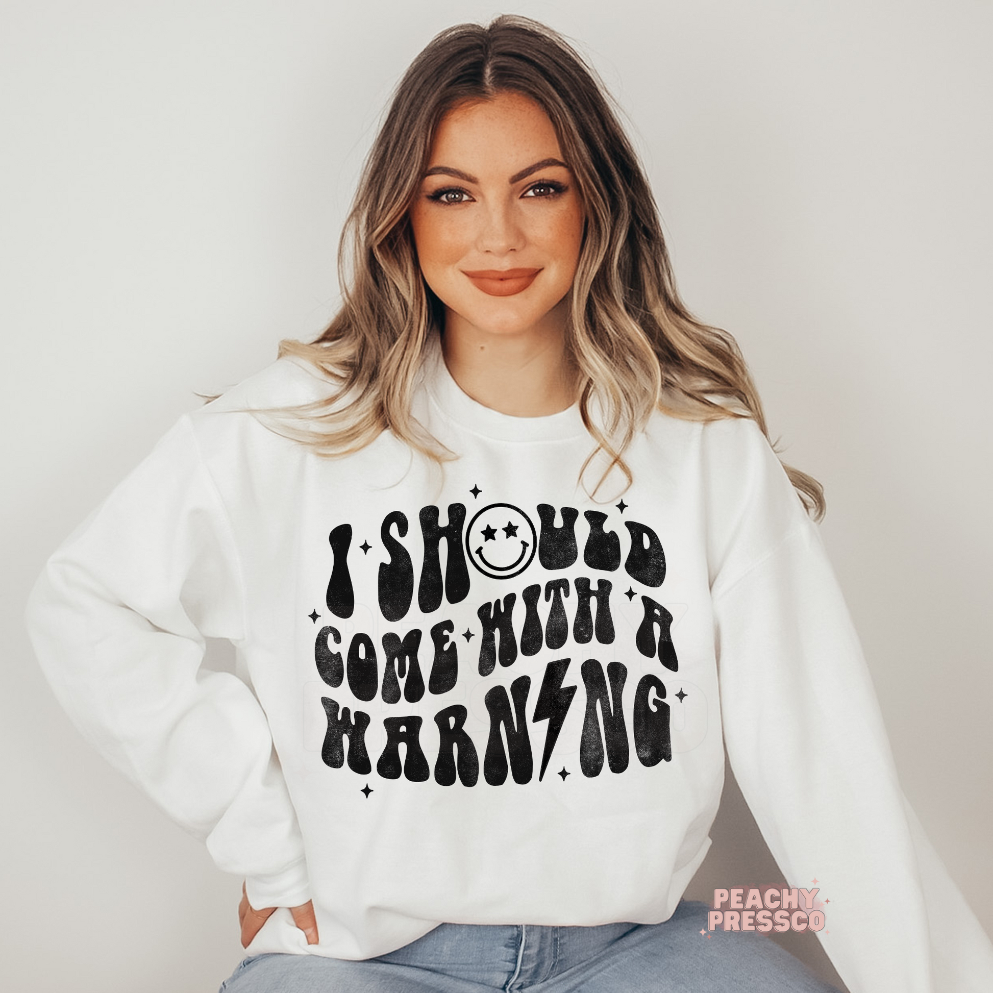Woman wearing a white crewneck sweatshirt featuring a retro "I Should Come With A Warning" graphic and smiley face.