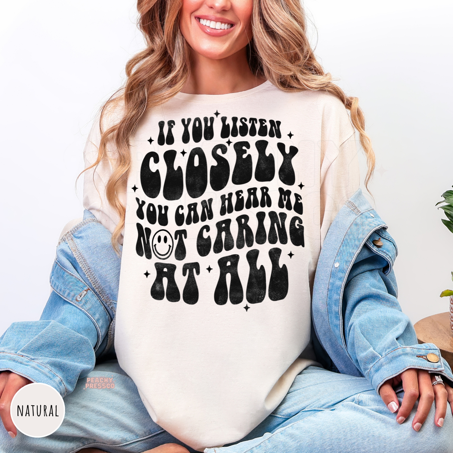 Woman wearing a natural shirt featuring the funny sarcastic quote "If You Listen Closely You Can Hear Me Not Caring at All" in distressed retro black typography.