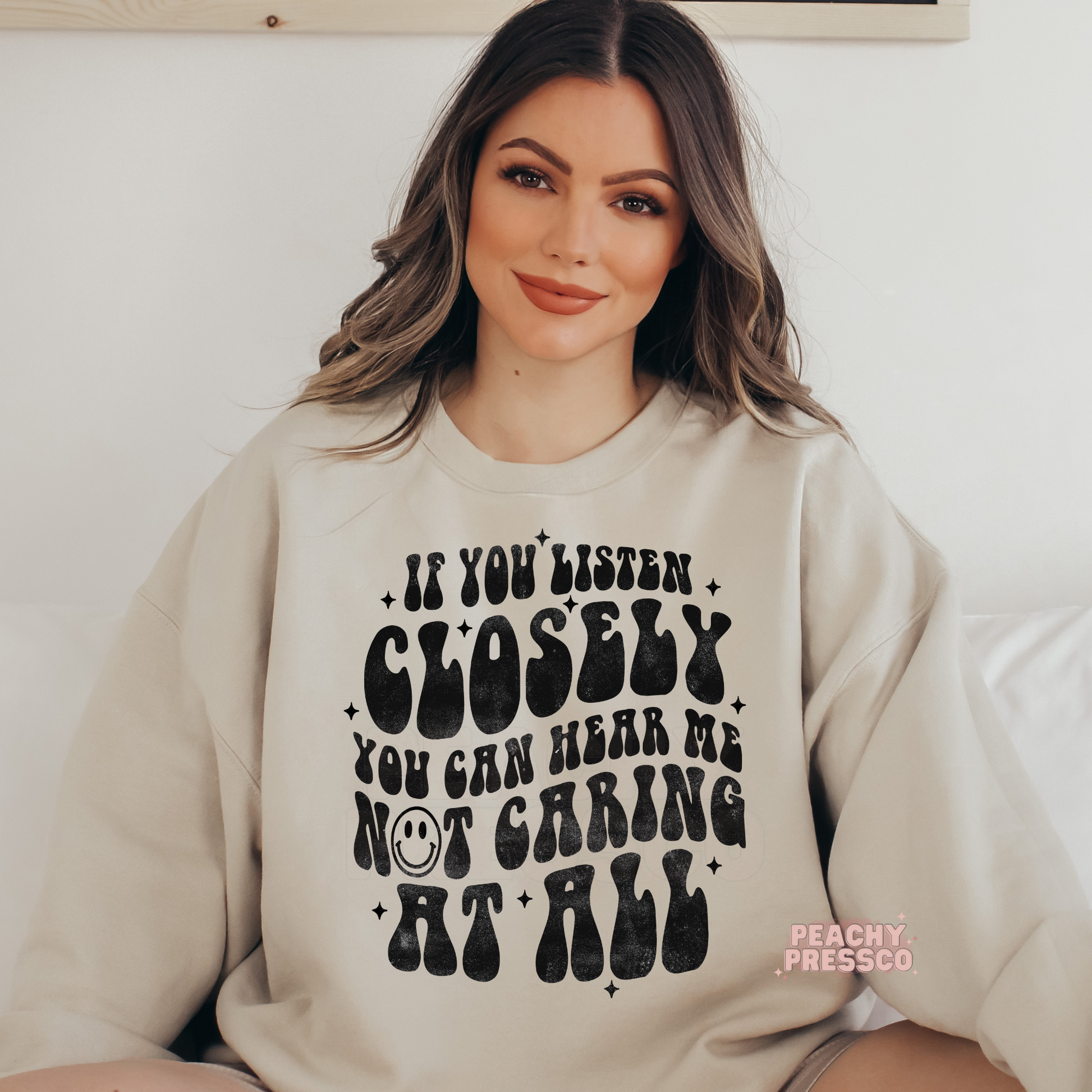 Sand beige sweatshirt displaying the funny quote "If You Listen Closely You Can Hear Me Not Caring at All" in oversized vintage black lettering.
