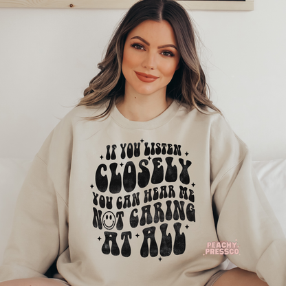 Sand beige sweatshirt displaying the funny quote "If You Listen Closely You Can Hear Me Not Caring at All" in oversized vintage black lettering.