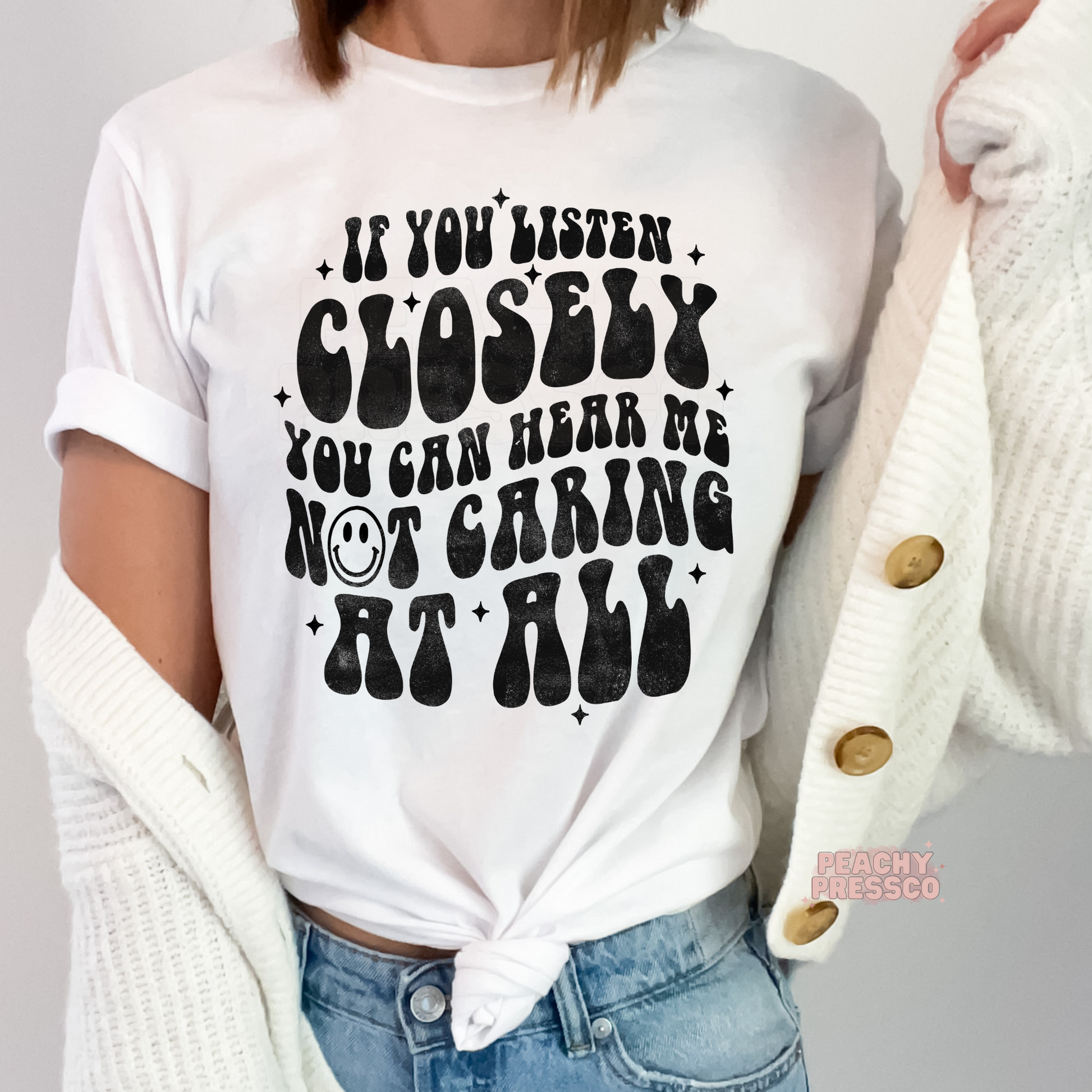 White graphic t-shirt with the humorous saying "If You Listen Closely You Can Hear Me Not Caring at All" in bold vintage black lettering with a smiley face accent.