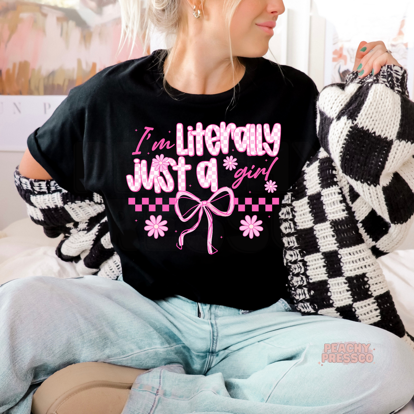 Woman wearing a black graphic t-shirt featuring the phrase "Literally Just a Girl" with pink coquette bow, checkered accents, and daisy flowers.