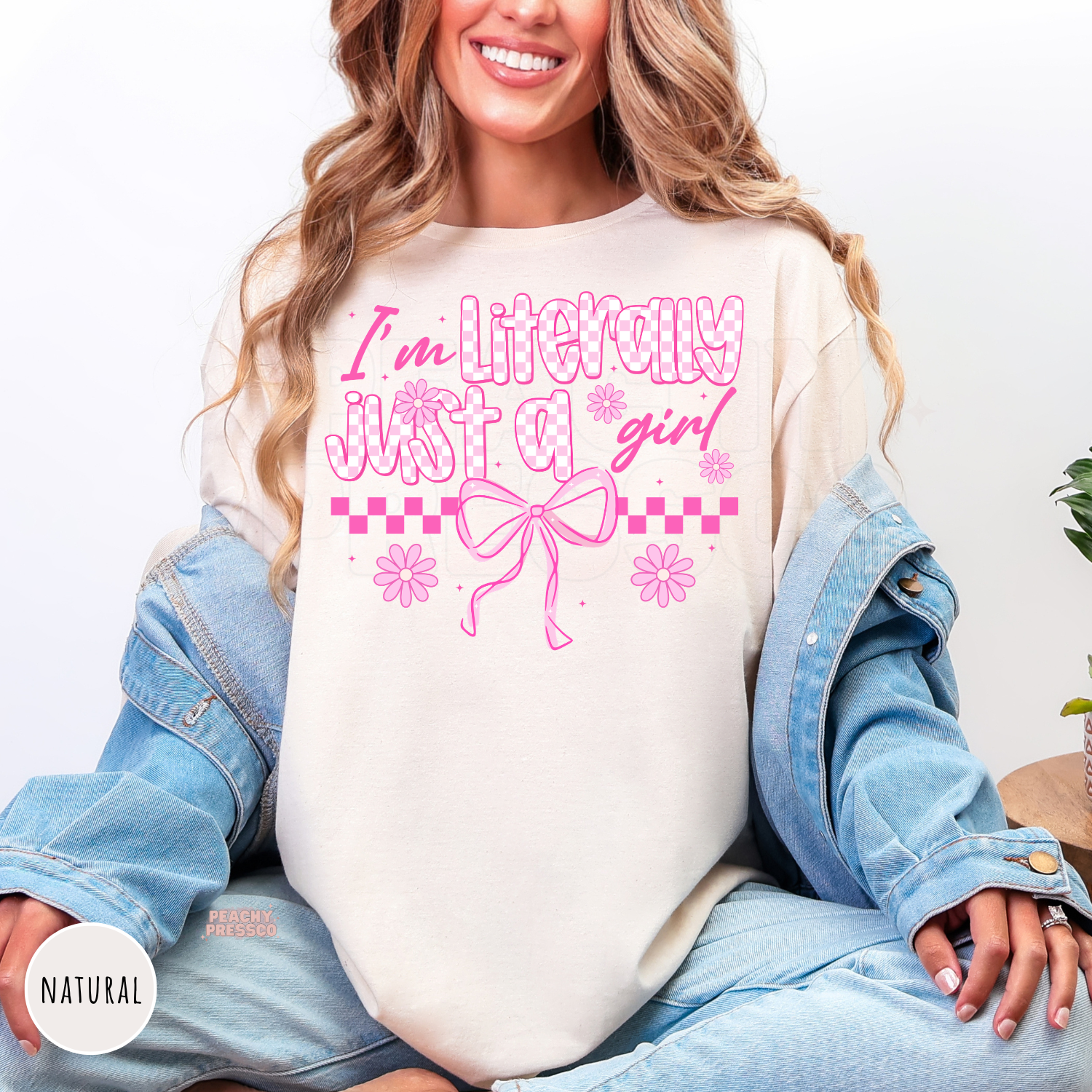 Woman wearing a natural colored shirt featuring a pink "I'm Literally Just a Girl" coquette design with bows, flowers, and checkerboard details.