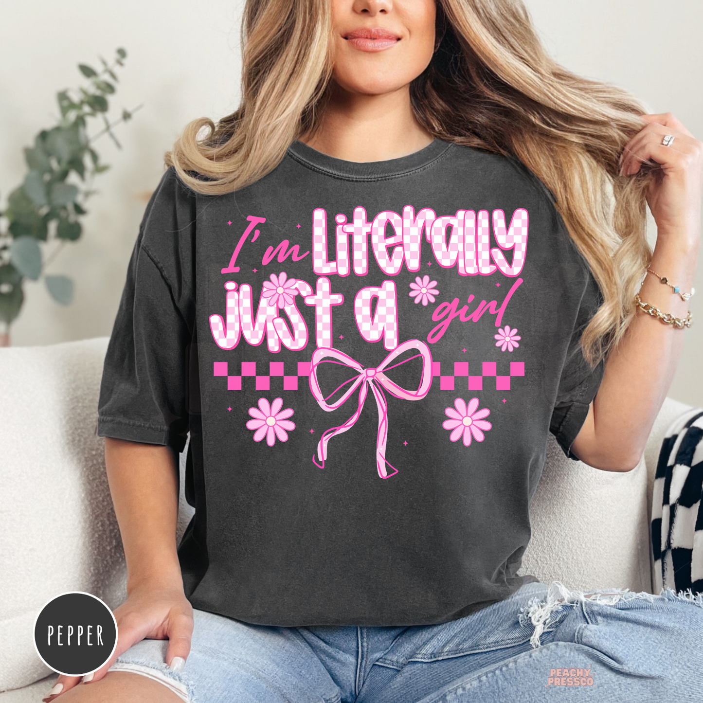 Woman wearing a pepper Comfort Colors t-shirt with a pink "I'm Literally Just a Girl" coquette graphic featuring bows, daisies, and checkered accents.