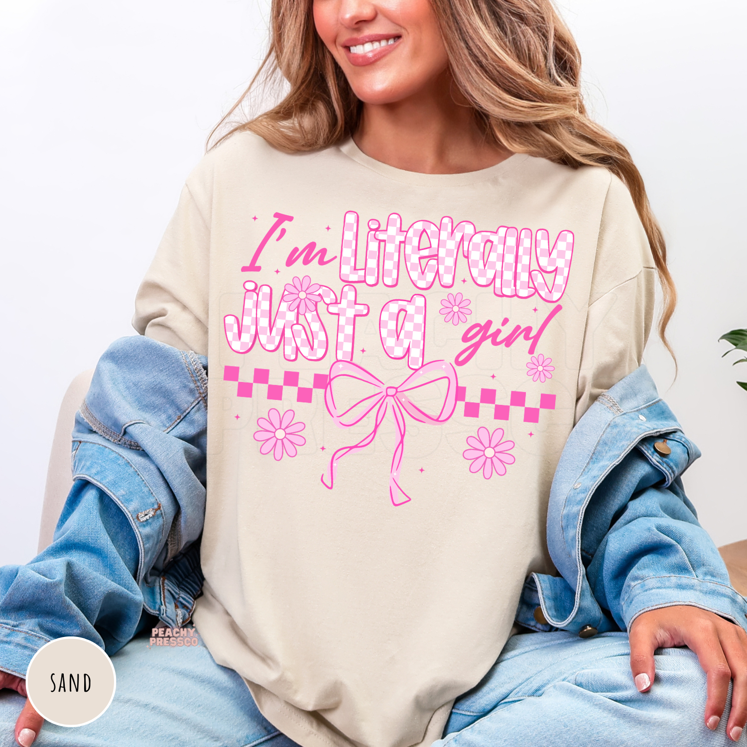 Woman wearing a sand crewneck sweatshirt featuring a pink coquette "I'm Literally Just a Girl" graphic with bows, flowers, and checkerboard details.
