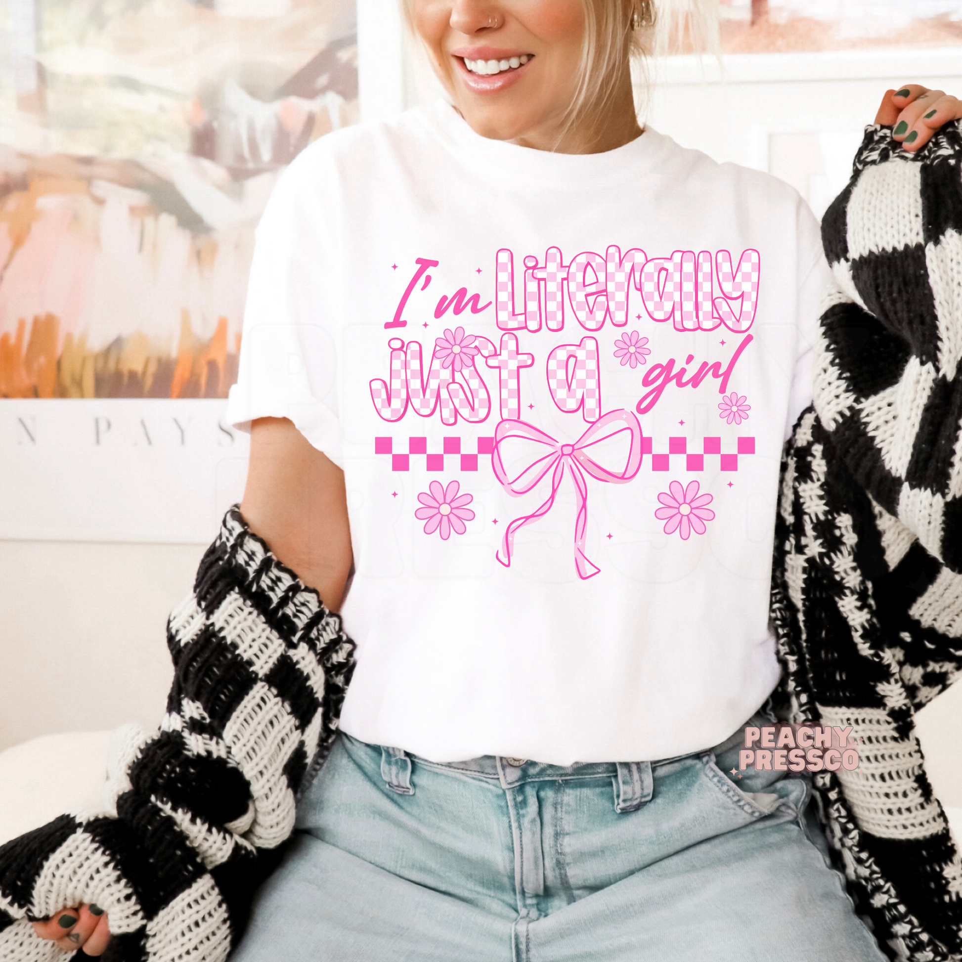 Woman wearing a white graphic t-shirt with a pink "I'm Literally Just a Girl" coquette design featuring a large bow, daisies, and checkerboard accents.