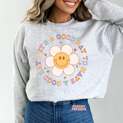 Ash grey sweatshirt with a retro smiley flower graphic and pastel typography featuring the uplifting quote "It's a Good Day to Have a Good Day."