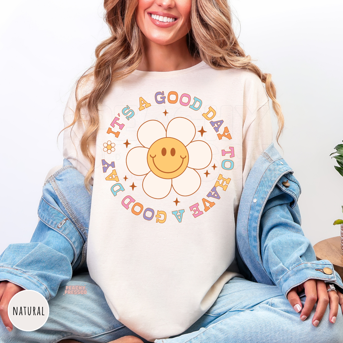 Natural shirt featuring a smiling daisy with pastel retro lettering that reads "It's a Good Day to Have a Good Day" in a cheerful vintage-inspired design.