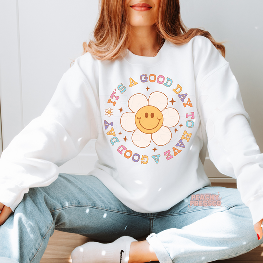 White sweatshirt featuring a smiling daisy surrounded by colorful retro lettering with the inspirational phrase "It's a Good Day to Have a Good Day."